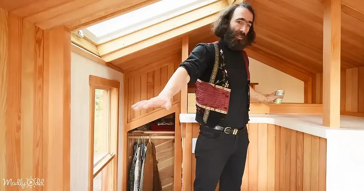 A man with a beard and floral vest stands in a wooden room, gesturing with one hand while holding a can in the other.