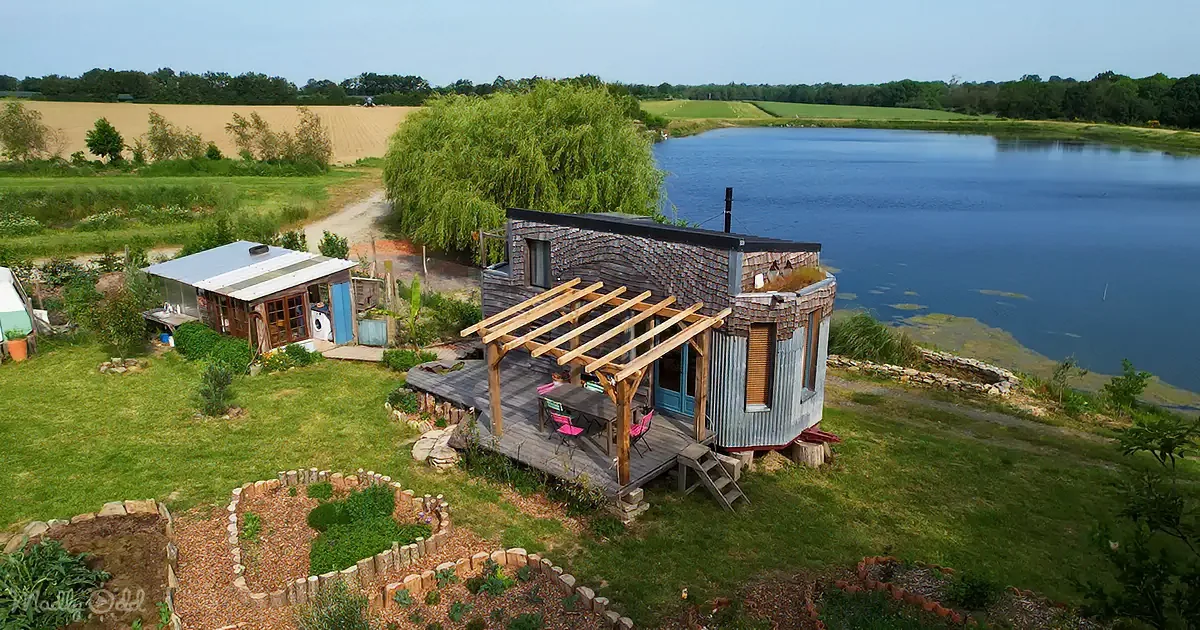 A rustic cabin with a wooden deck and a small garden sits by a serene lake surrounded by lush greenery and farmland.