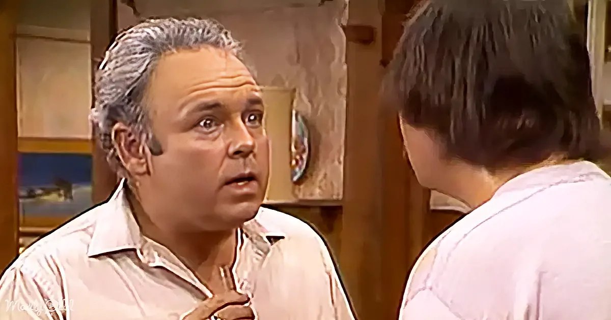 A man in a white shirt gestures with his hand while talking to another person in a room with wooden decor.