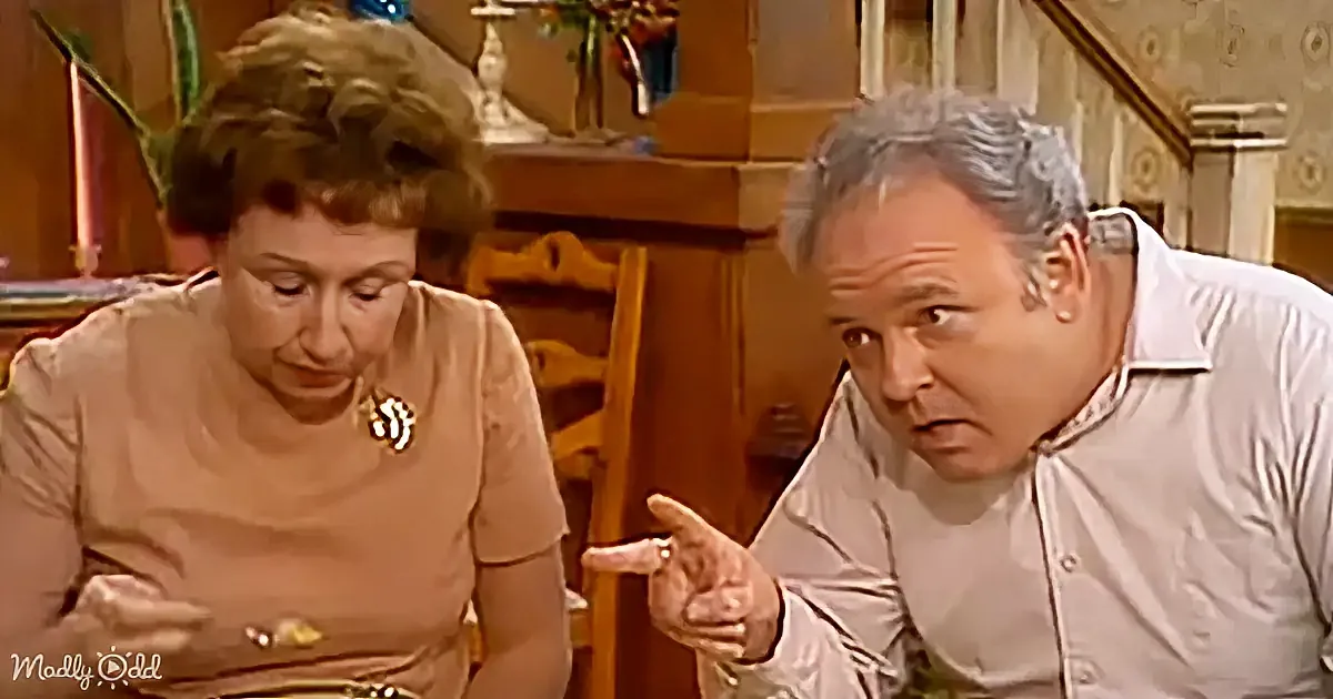 A man in a white shirt gestures while talking to a woman in a brown dress in a warmly lit room.