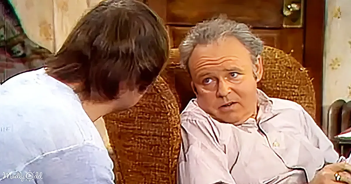A man in a white shirt sits in a wicker chair, looking concerned, while another person leans in closely.