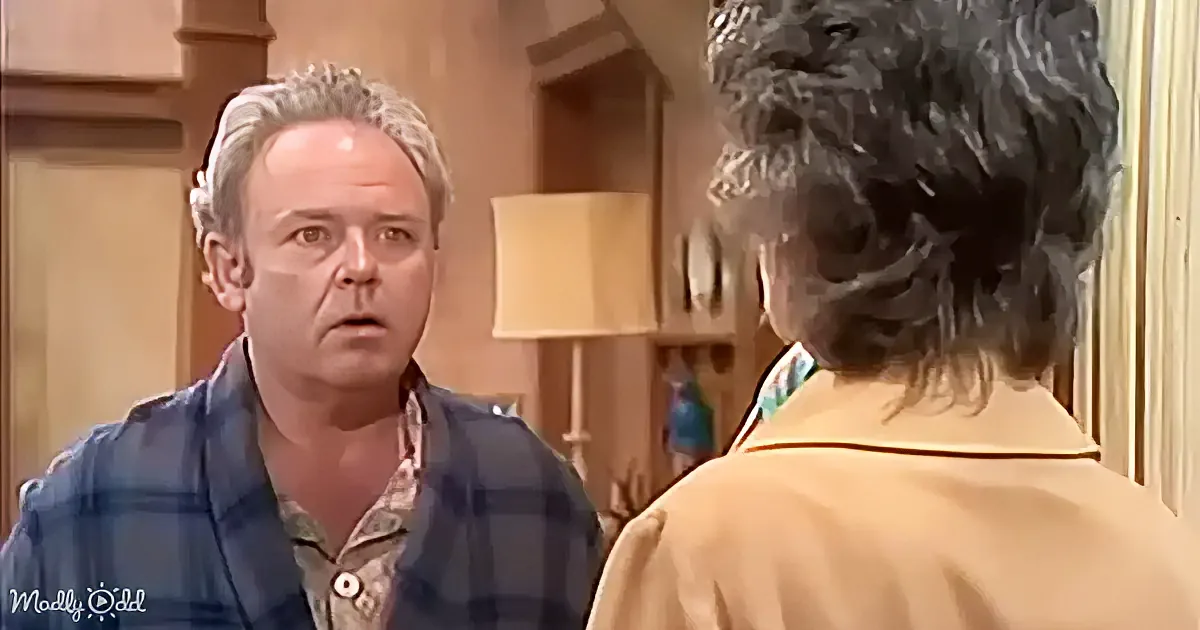 A man in a plaid shirt looks surprised, facing another person in a tan jacket.