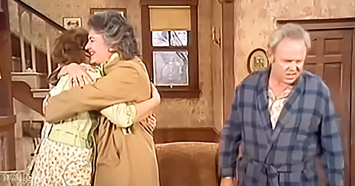 A woman in a tan coat hugs another woman in a green dress, while a man in a plaid robe looks on.