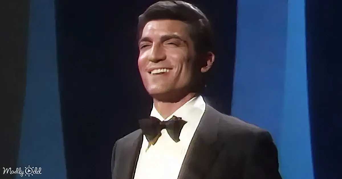 A man in a suit and bow tie smiles warmly, set against a dark blue background.