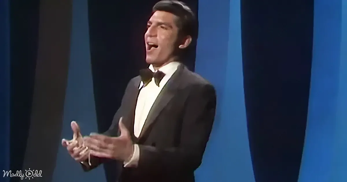 A man in a tuxedo is singing with his hands outstretched on stage.