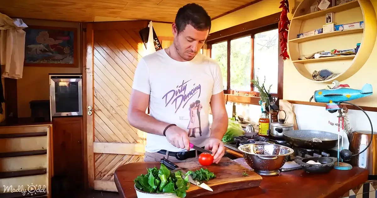 A man in a "Dirty Dancing" t-shirt is chopping vegetables on a wooden cutting board in a cozy kitchen.