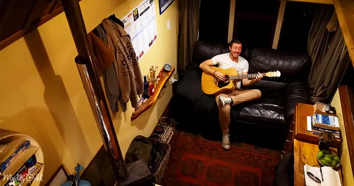 A man sits on a black leather couch playing an acoustic guitar in a cozy room with a red rug and various items around.