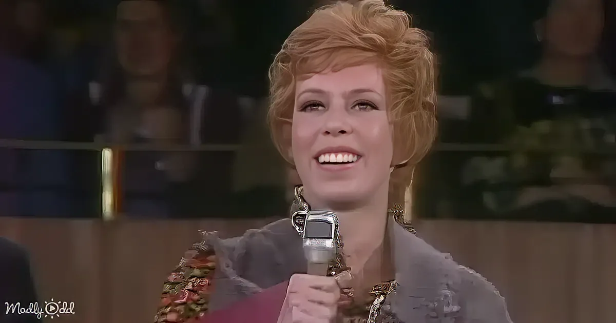 A smiling woman with short blonde hair holds a microphone, wearing a patterned top and earrings.