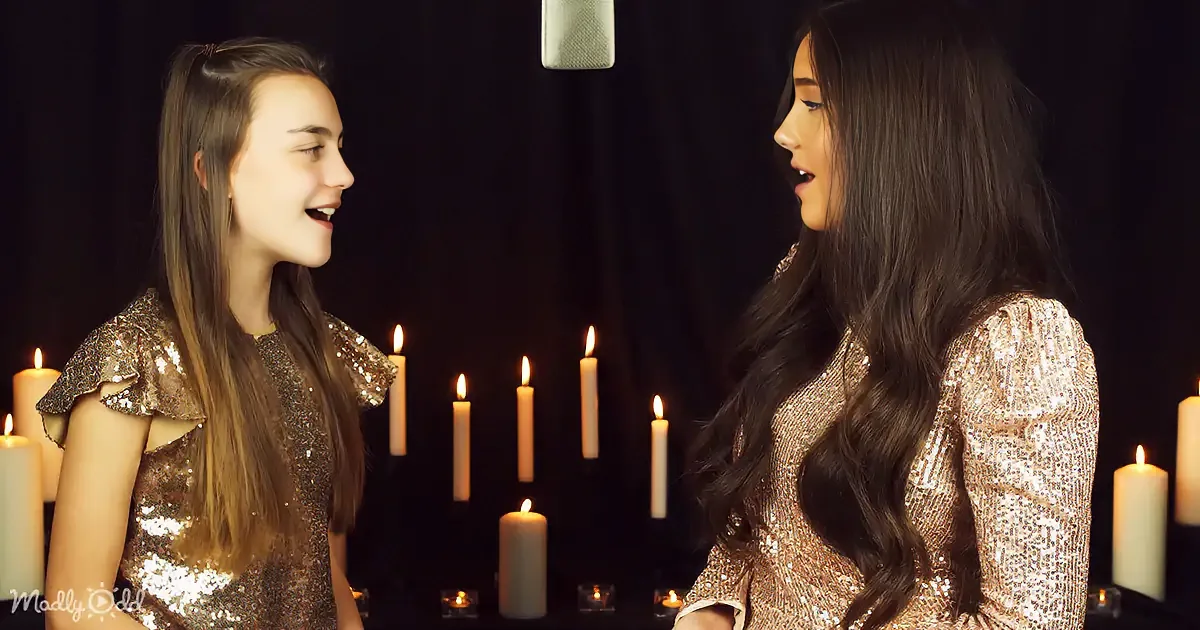 Two women in sparkly dresses sing in front of lit candles against a dark background.