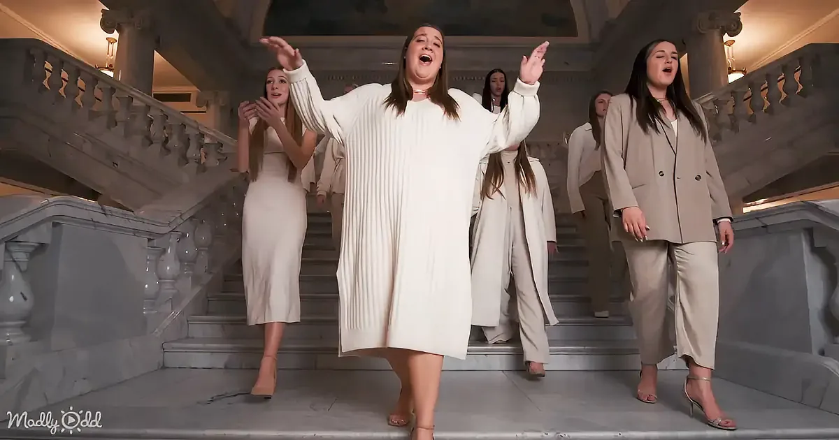 A woman in a white dress stands on a grand staircase, arms raised, singing passionately.