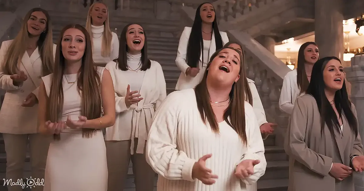 A woman in white leads a choir in song, surrounded by others in formal attire, in a grand, ornate setting.