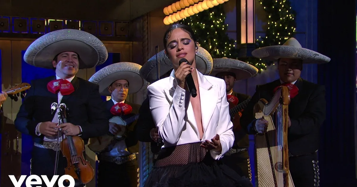 A woman in a white jacket sings into a microphone, surrounded by musicians in sombreros.