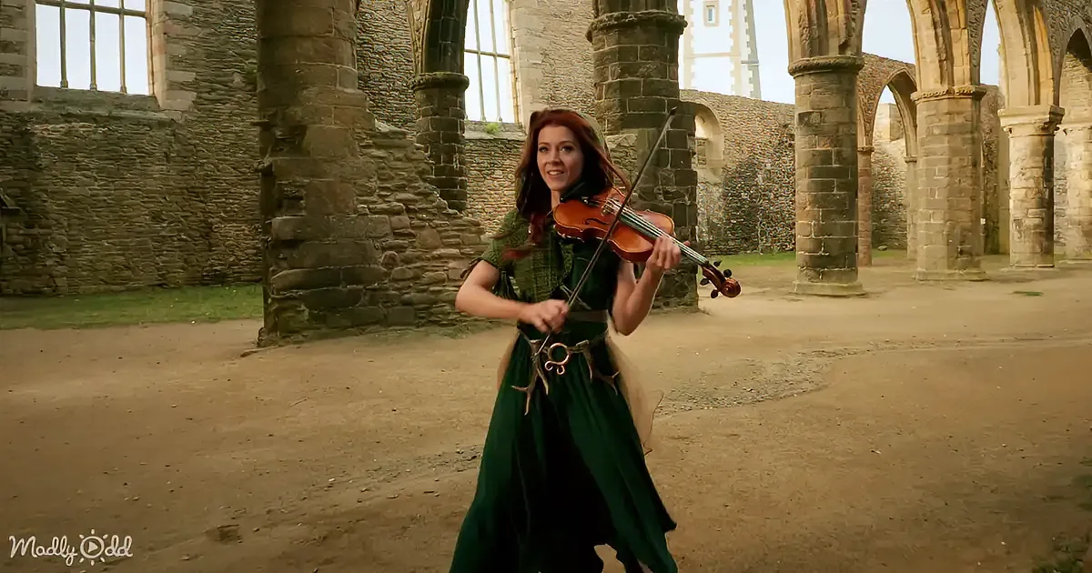 A woman in a green dress plays a violin in an ancient stone ruin.