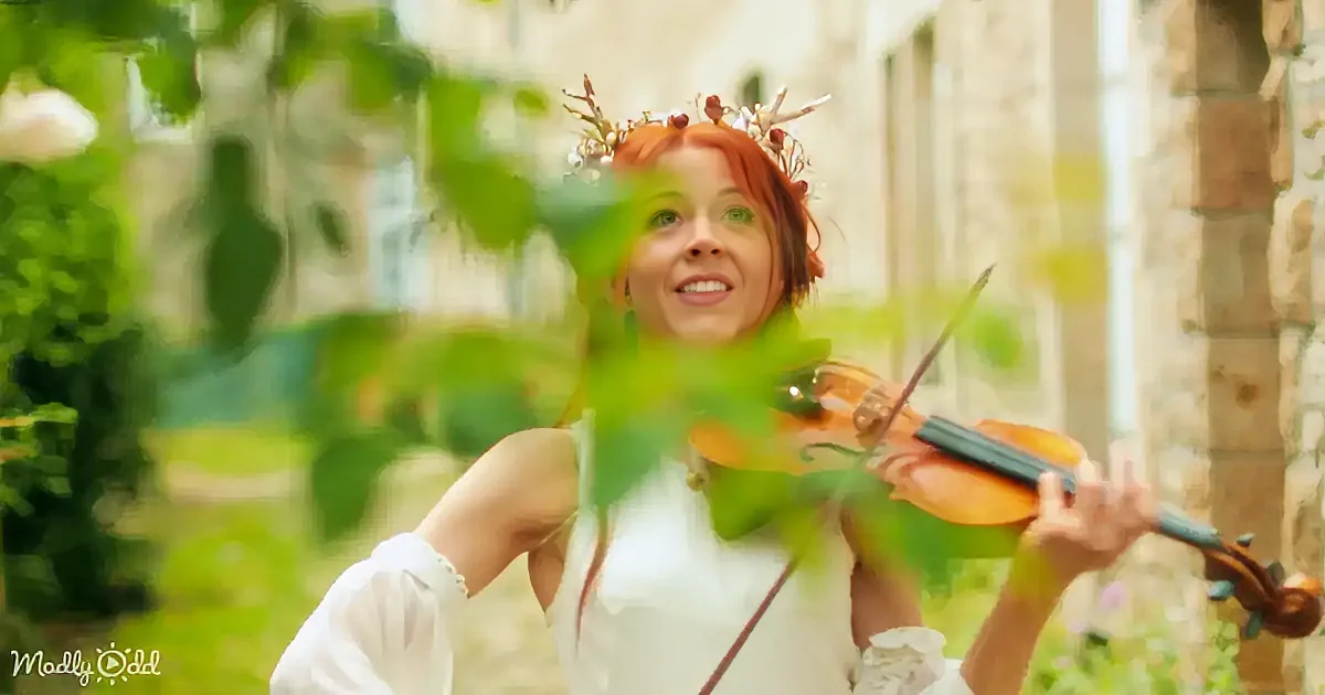 A woman in a white dress plays a violin outdoors, adorned with a crown of twigs and berries.