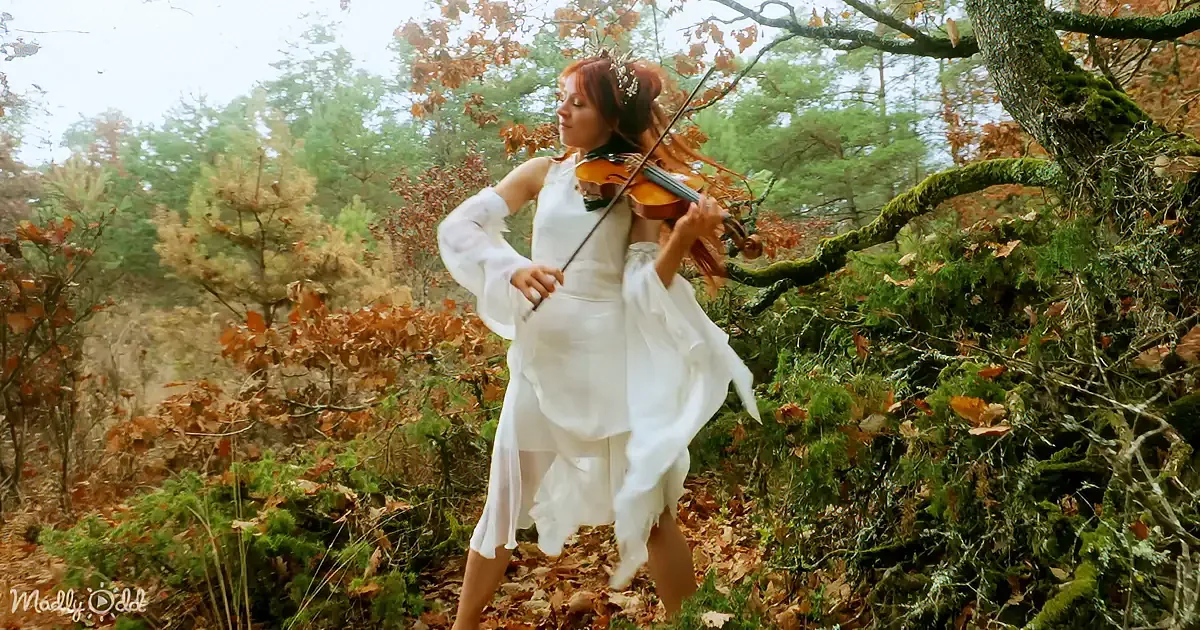 A woman in a white dress plays a violin amidst autumn leaves and mossy trees.