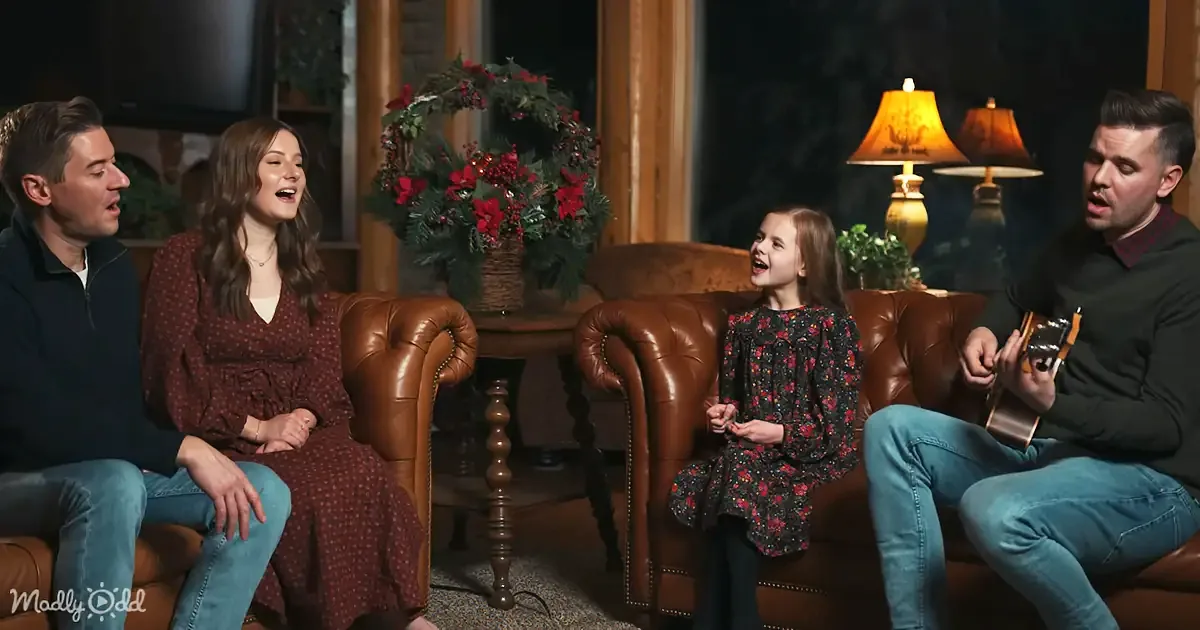 A family of four, including a child, sits in a cozy living room, singing and playing guitar, with a festive wreath and...