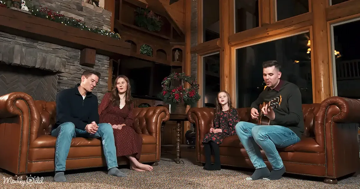 A family of four sits in a cozy living room, with a man playing guitar and a young girl singing.