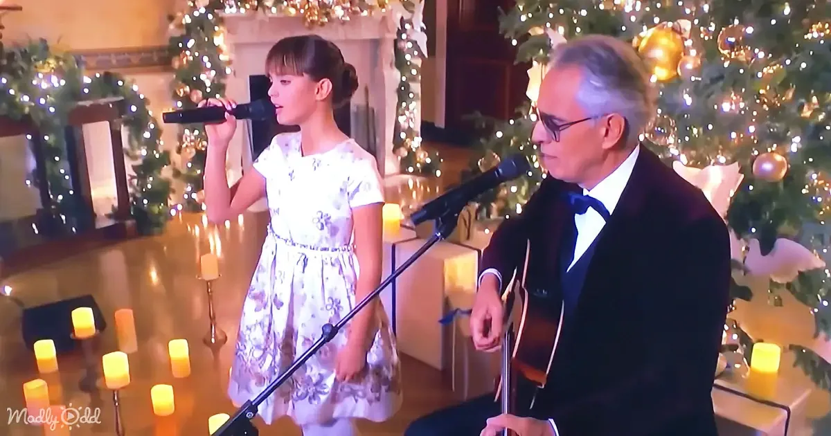 A young girl in a floral dress sings into a microphone, accompanied by a man in a suit playing guitar, both in front of...