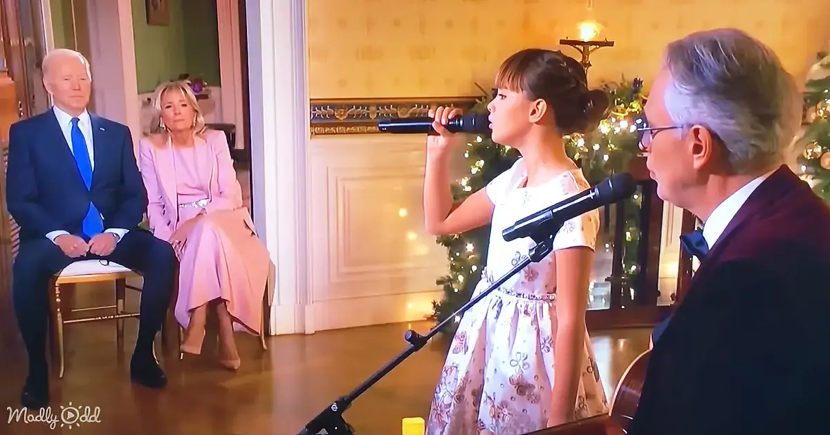 A young girl in a floral dress sings into a microphone, surrounded by adults in formal attire, with a Christmas tree in...