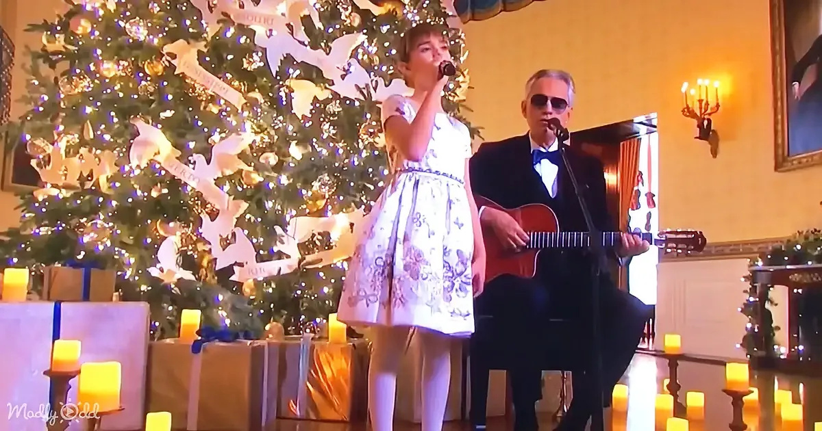 A young girl sings into a microphone, accompanied by a man playing guitar, in front of a decorated Christmas tree.