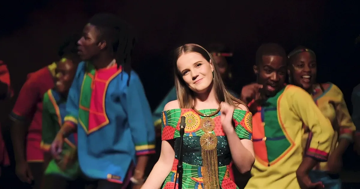 A woman in a colorful dress sings into a microphone, surrounded by a choir in vibrant attire.