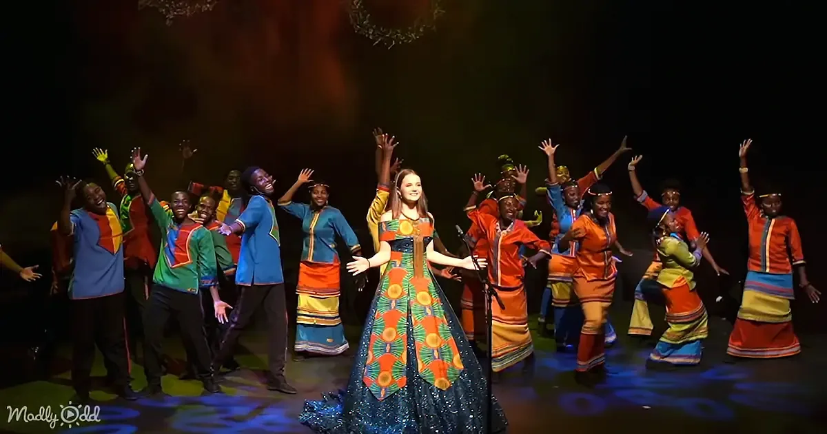 A woman in a colorful dress stands center stage, surrounded by a group of performers in vibrant costumes, all raising...