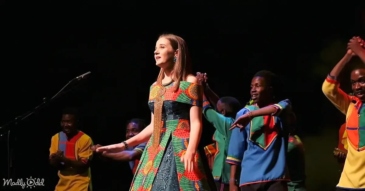 A woman in a colorful dress stands on stage with a group of performers in vibrant outfits.