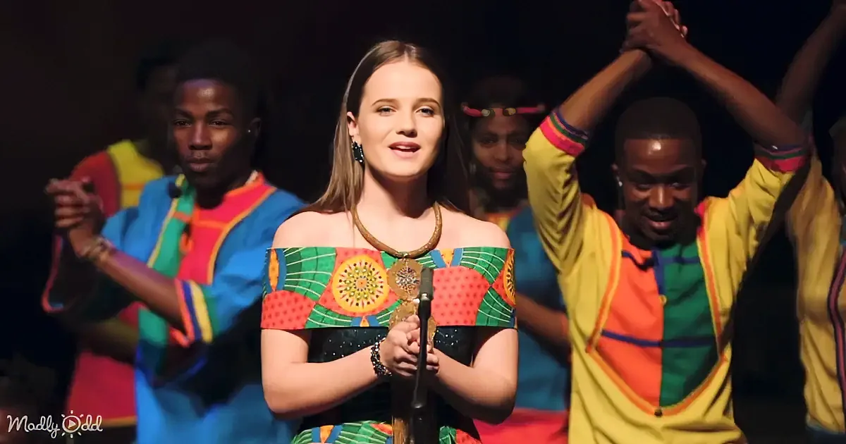 A woman in a colorful, patterned dress sings into a microphone, surrounded by dancers in vibrant outfits.