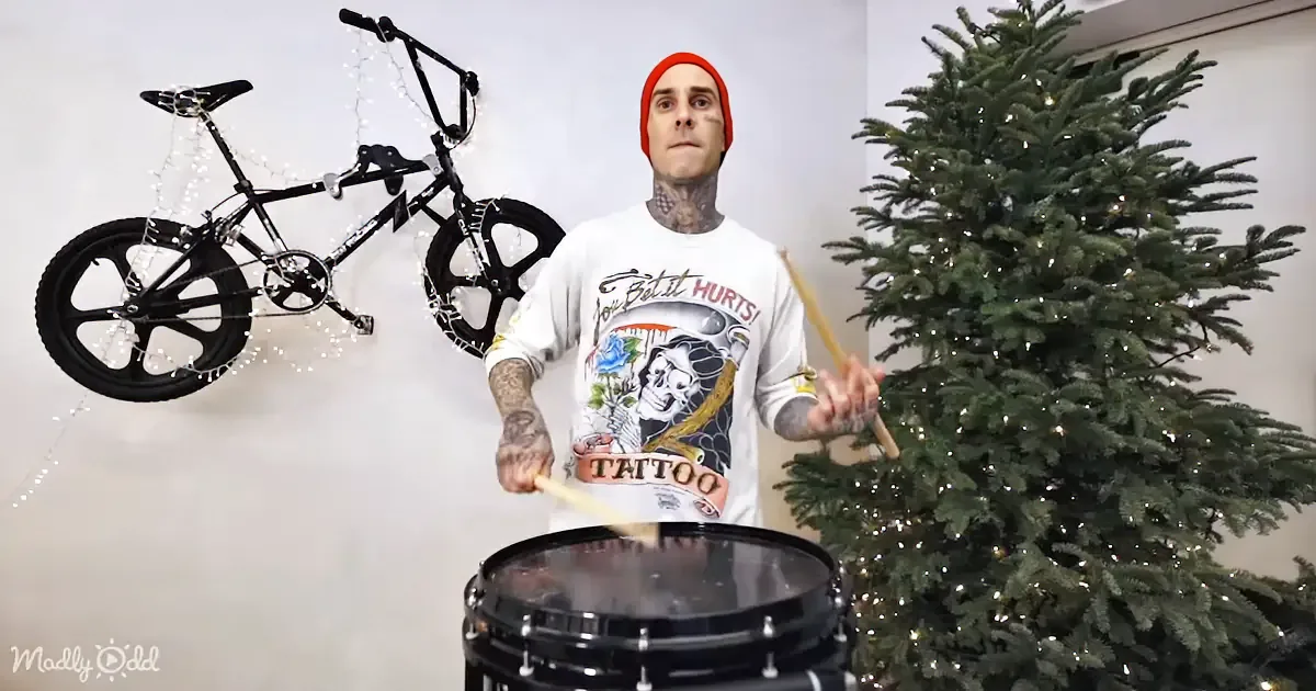 A man with tattoos plays a drum set next to a Christmas tree, with a bicycle decorated with lights in the background.