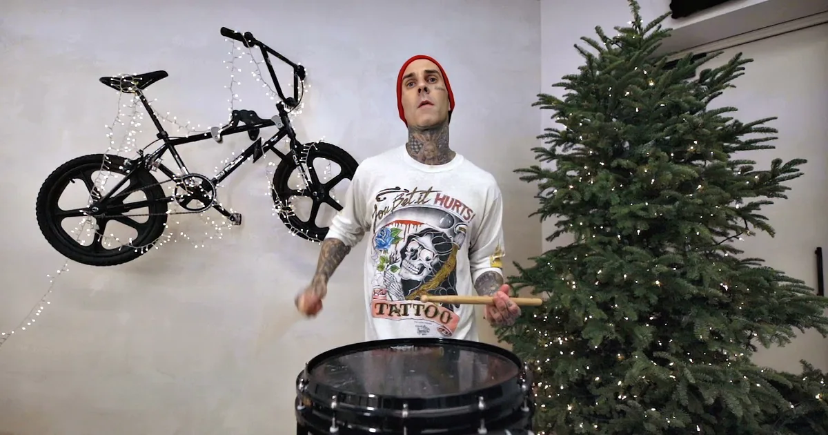 A man with tattoos plays drums near a decorated Christmas tree, with a bicycle adorned with lights hanging on the wall.