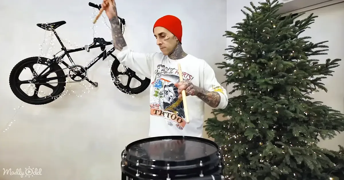 A tattooed man in a red beanie plays a drum with a bicycle and Christmas tree in the background.