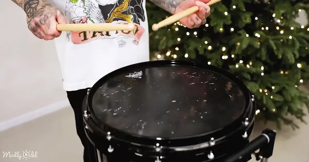 A tattooed person holds drumsticks above a black drum, with a Christmas tree in the background.