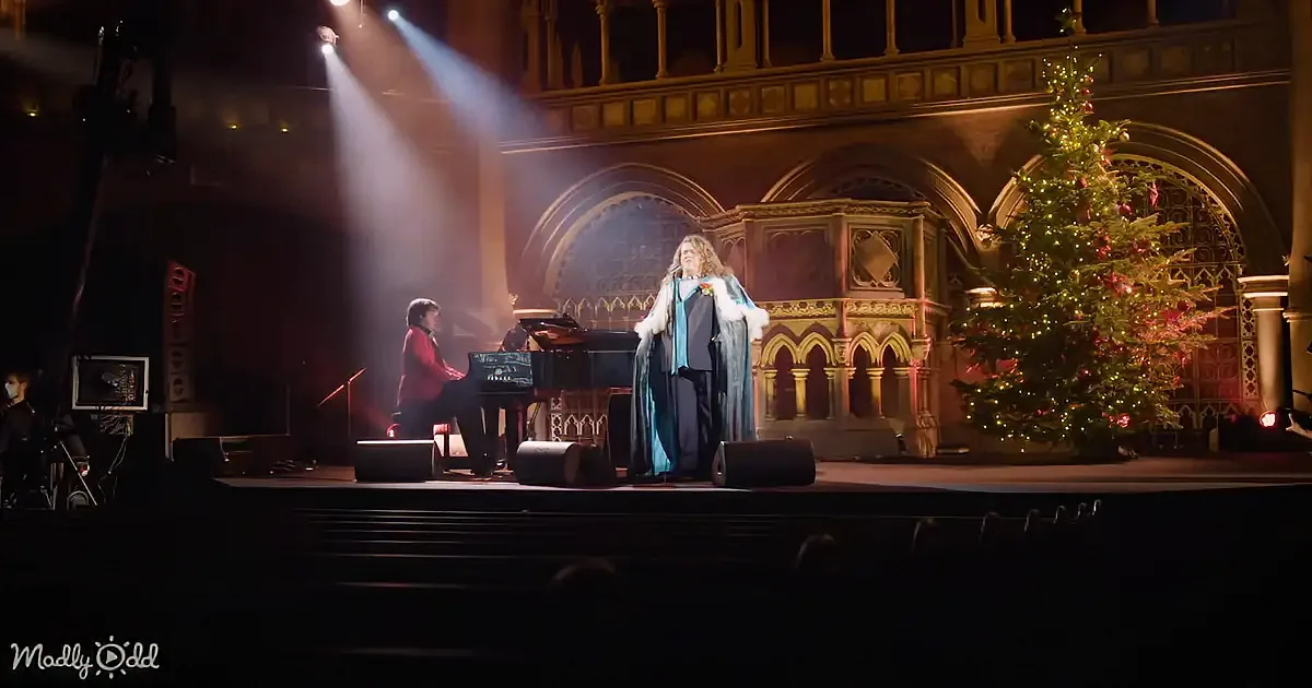 A performer in a blue robe sings on stage with a pianist, a Christmas tree, and ornate architecture in the background.