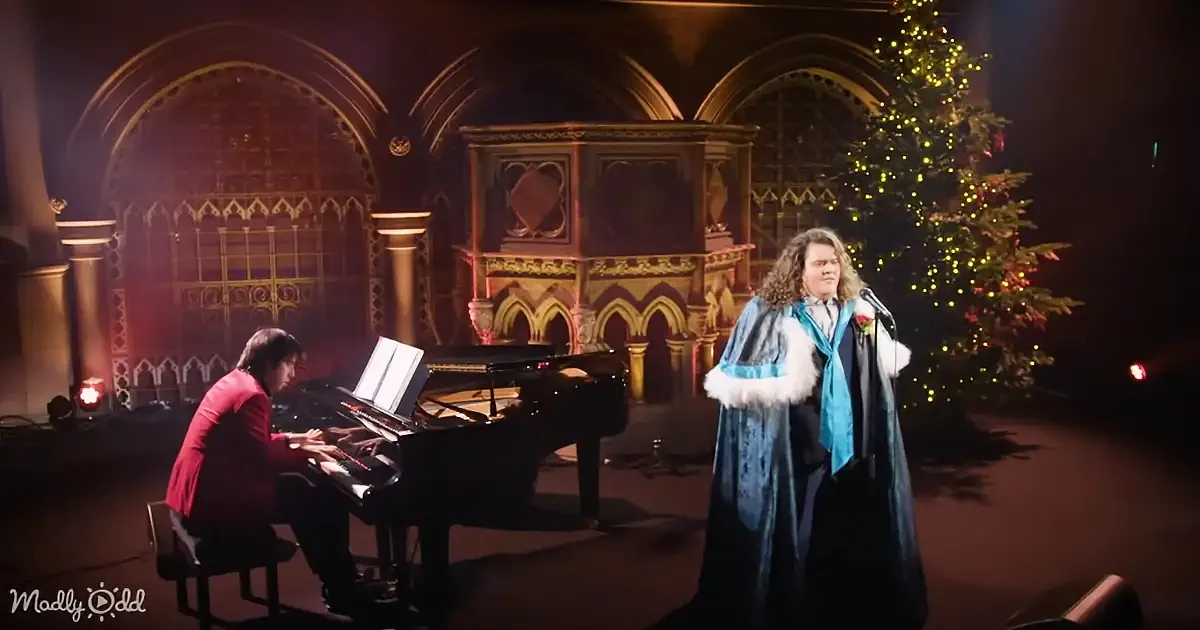 A person in a blue robe sings into a microphone while another plays piano in a grand, ornate setting with a Christmas tree.