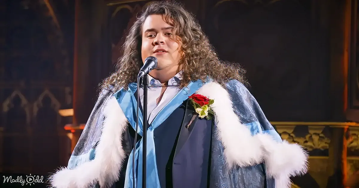 A person in a blue and white fur-trimmed robe stands at a microphone, adorned with a red rose, in a dimly lit, ornate...
