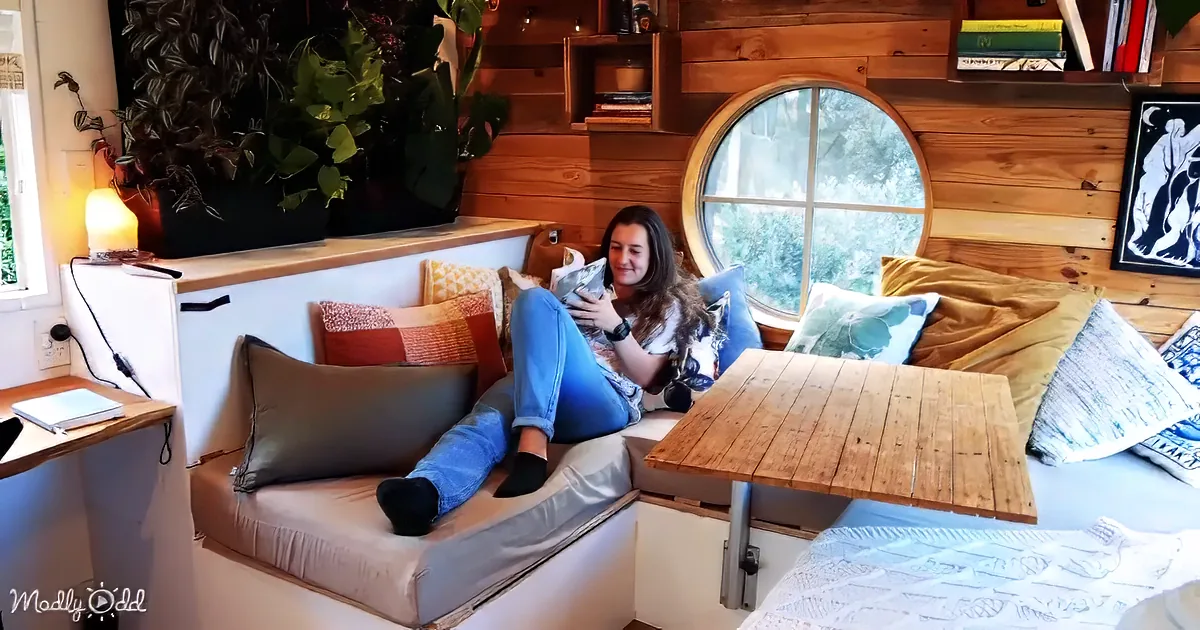 A woman sits on a cozy, cushioned bench reading in a rustic, well-lit room with wooden walls and a large circular window.