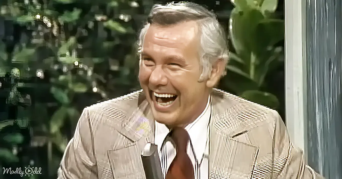 A man in a patterned suit jacket and tie is laughing heartily, with greenery in the background.