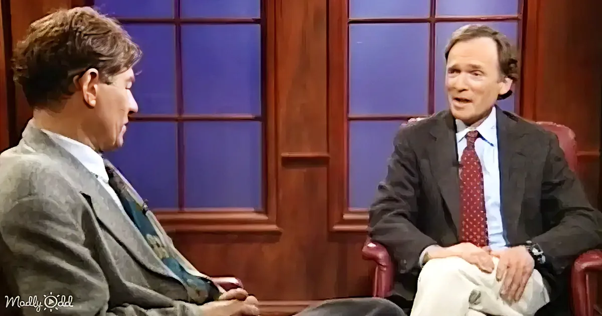Two men in suits are seated, engaged in a conversation on a talk show set with a wooden backdrop.