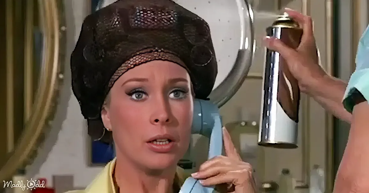 A woman in a yellow top and black headpiece is holding a phone, looking surprised, while someone sprays something from a can.