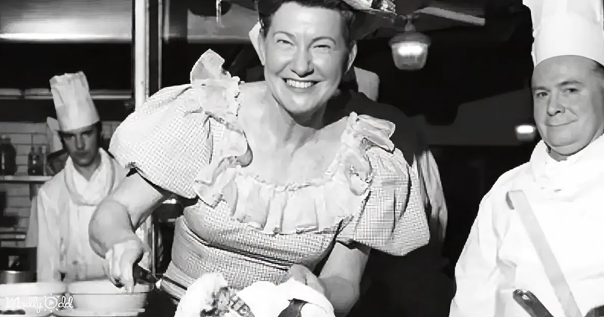 A smiling woman in a checkered dress holds a bag, with two chefs in the background.