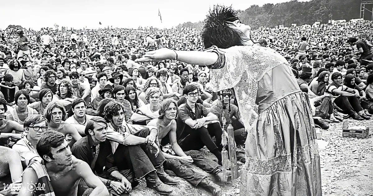 A woman in a dress stands in front of a large crowd, gesturing upwards, with a man in the foreground looking on.