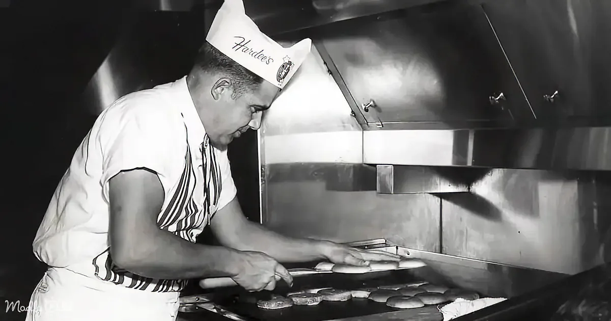 A chef in a Hardee's uniform cooks burgers on a grill in a kitchen.