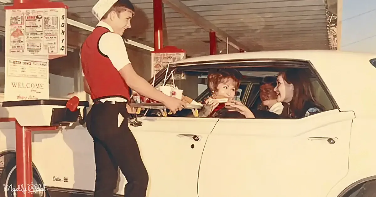 A server in a red vest hands food to a woman in a car at a drive-in.