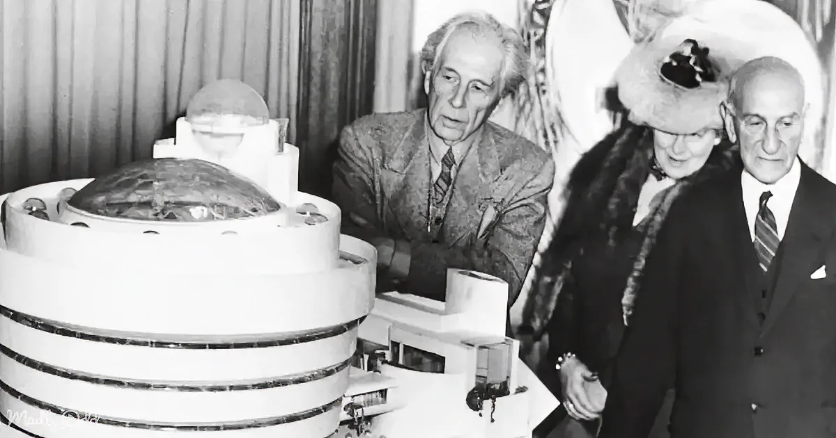 Three formally dressed individuals examine a futuristic, circular device with a dome on top.