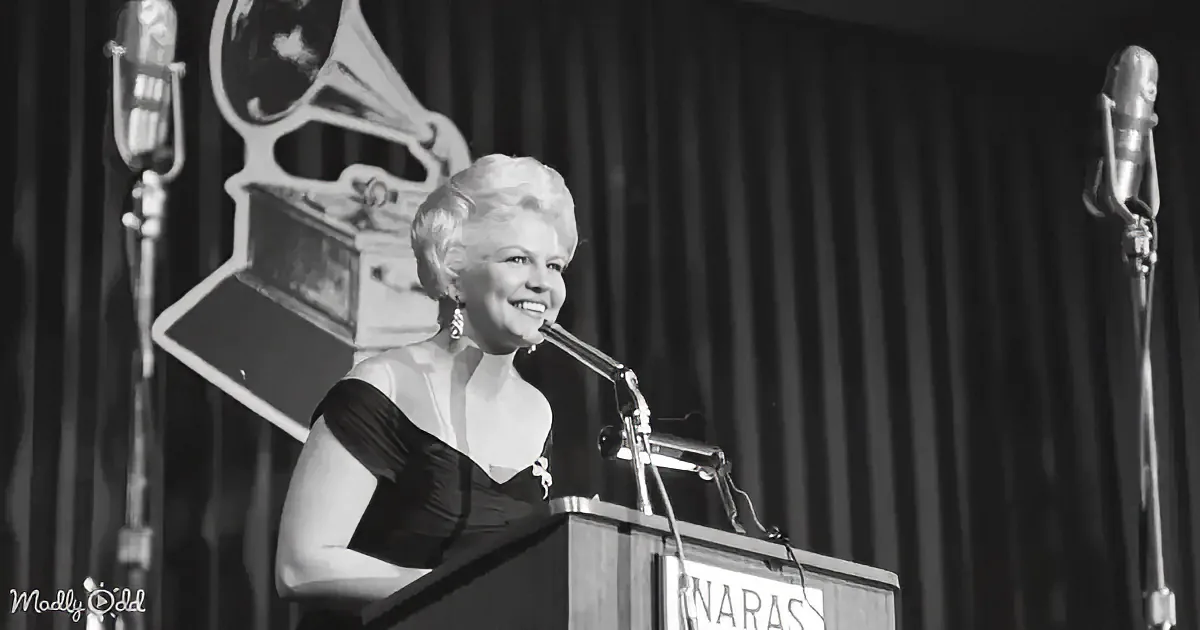 A woman in a black dress smiles at a podium with a microphone, surrounded by vintage microphones and a gramophone.