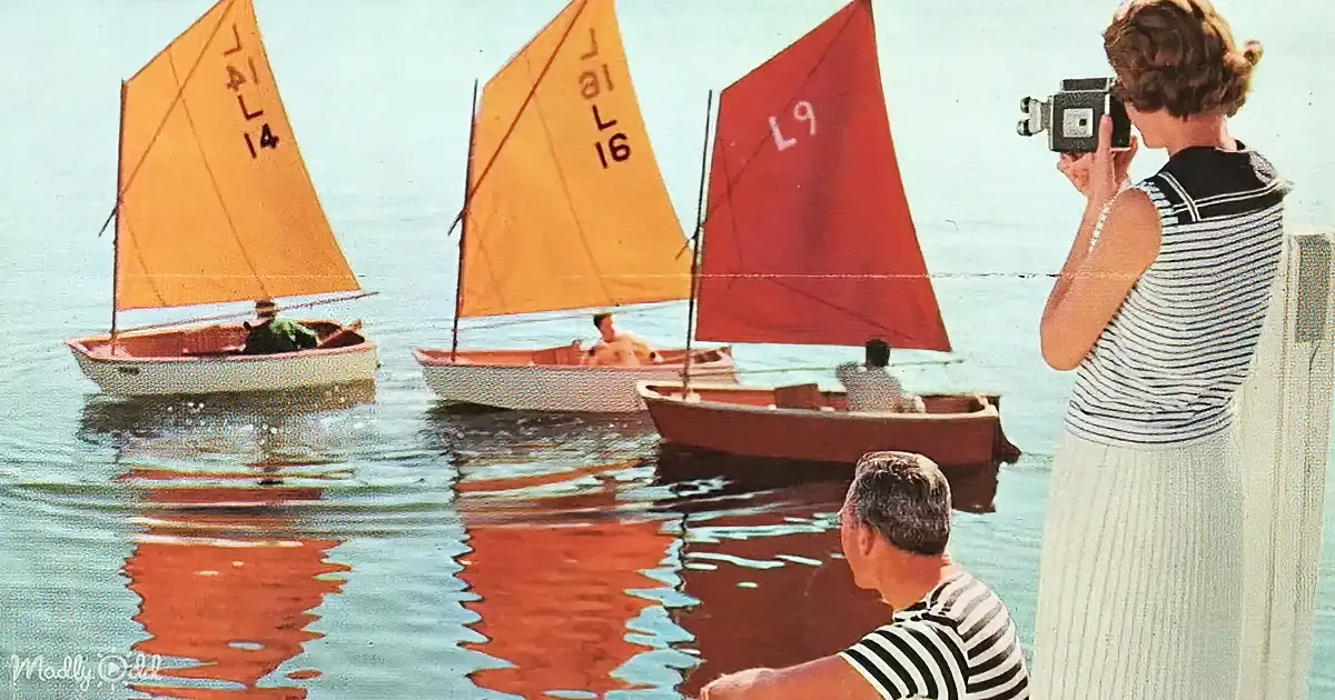 A woman films sailboats with numbered sails on a calm lake, while a man watches nearby.