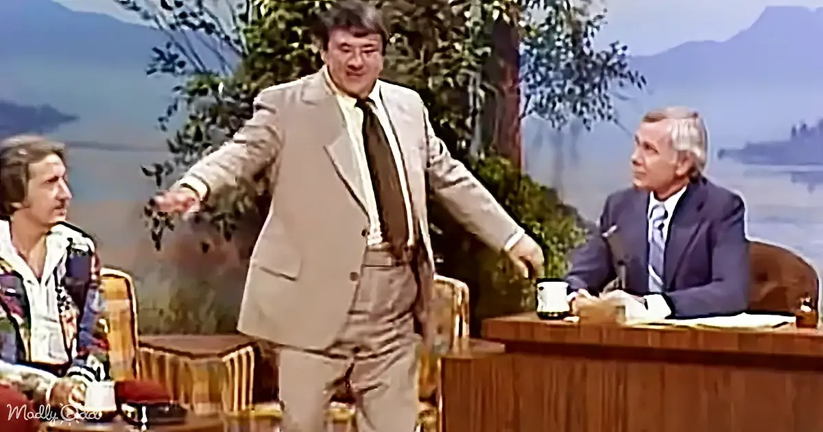A man in a tan suit gestures dramatically on a talk show set with a scenic backdrop.
