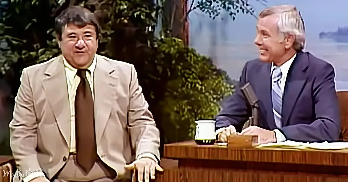 Steve Martin in a beige suit sits on a talk show set, smiling, with a host in a blue suit beside him.