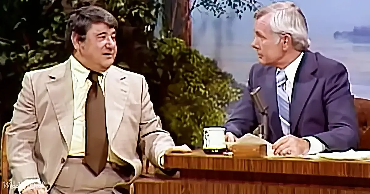 Two men in suits are seated at a desk, one in a beige suit and the other in a dark suit, engaged in conversation.