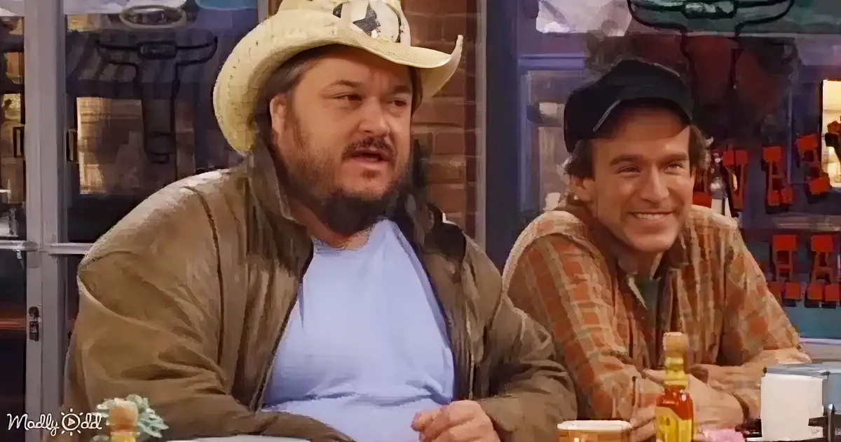 A man in a cowboy hat and brown jacket sits at a table, while another man in a plaid shirt smiles beside him.